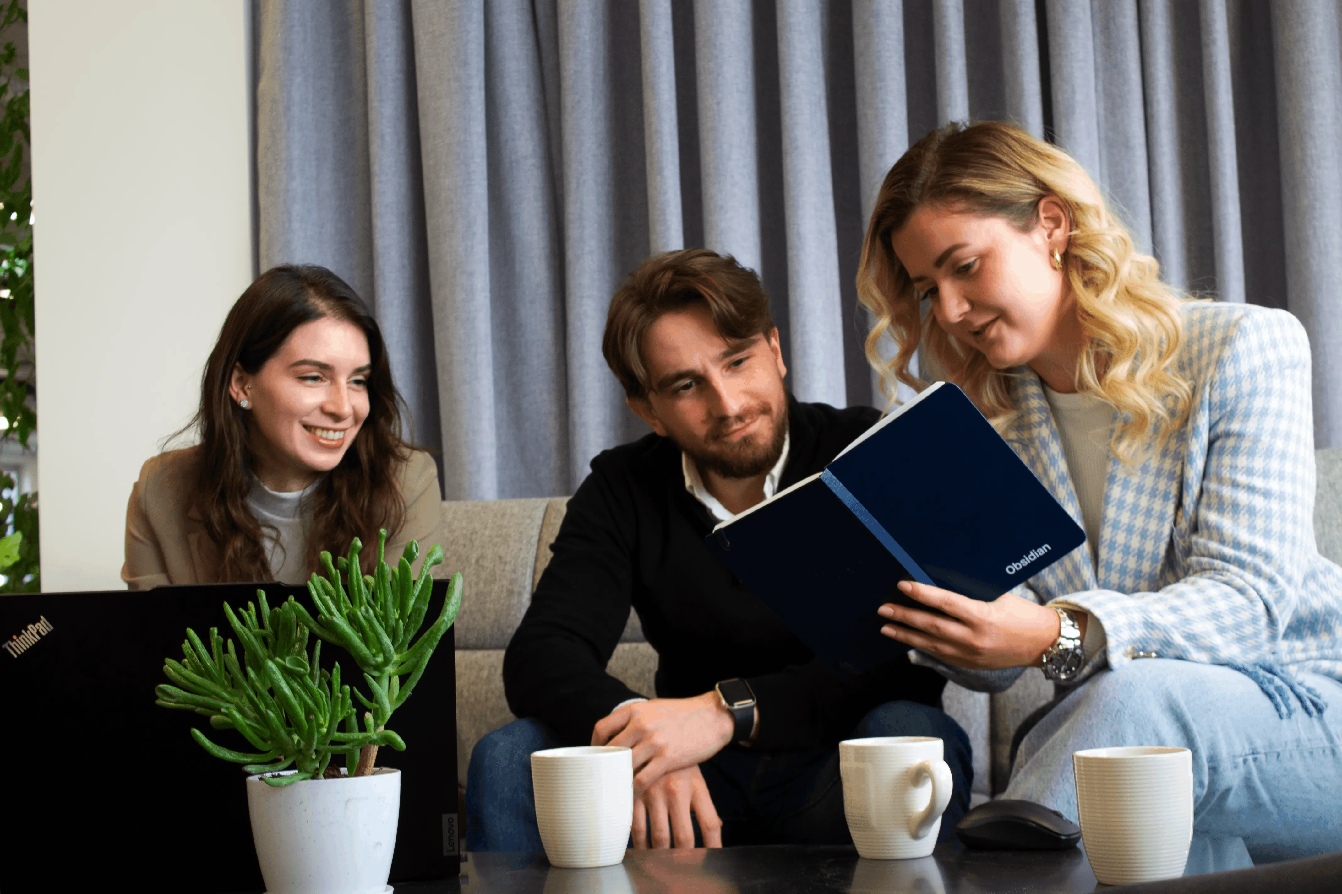 Three colleagues sitting on a couch, looking at a notebook, with a laptop and coffee mugs on the table.