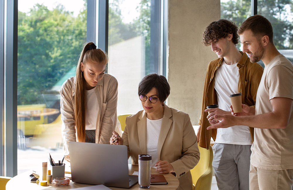 Four colleagues gathered around a laptop in a bright office, discussing work while holding coffee cups.