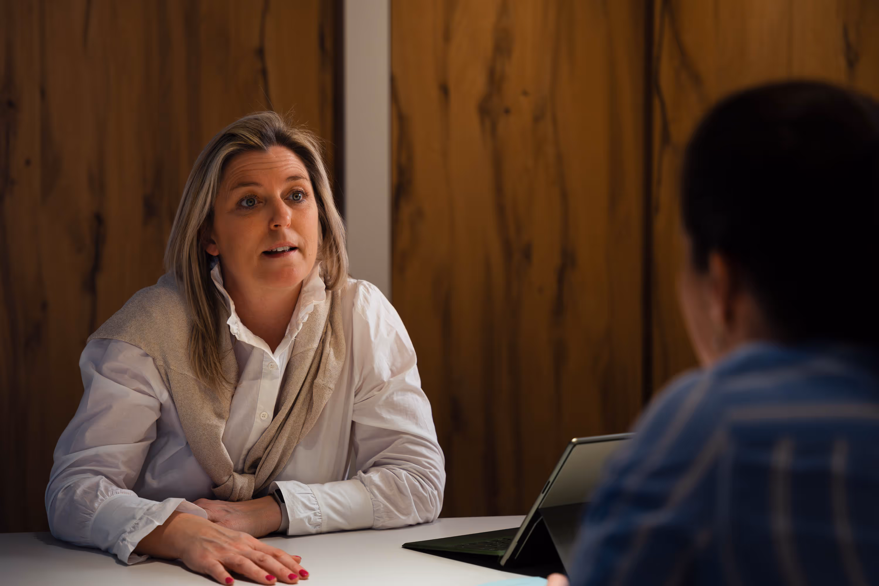 Woman wearing a white shirt and beige sweater sitting at a table engaged in conversation with another person.