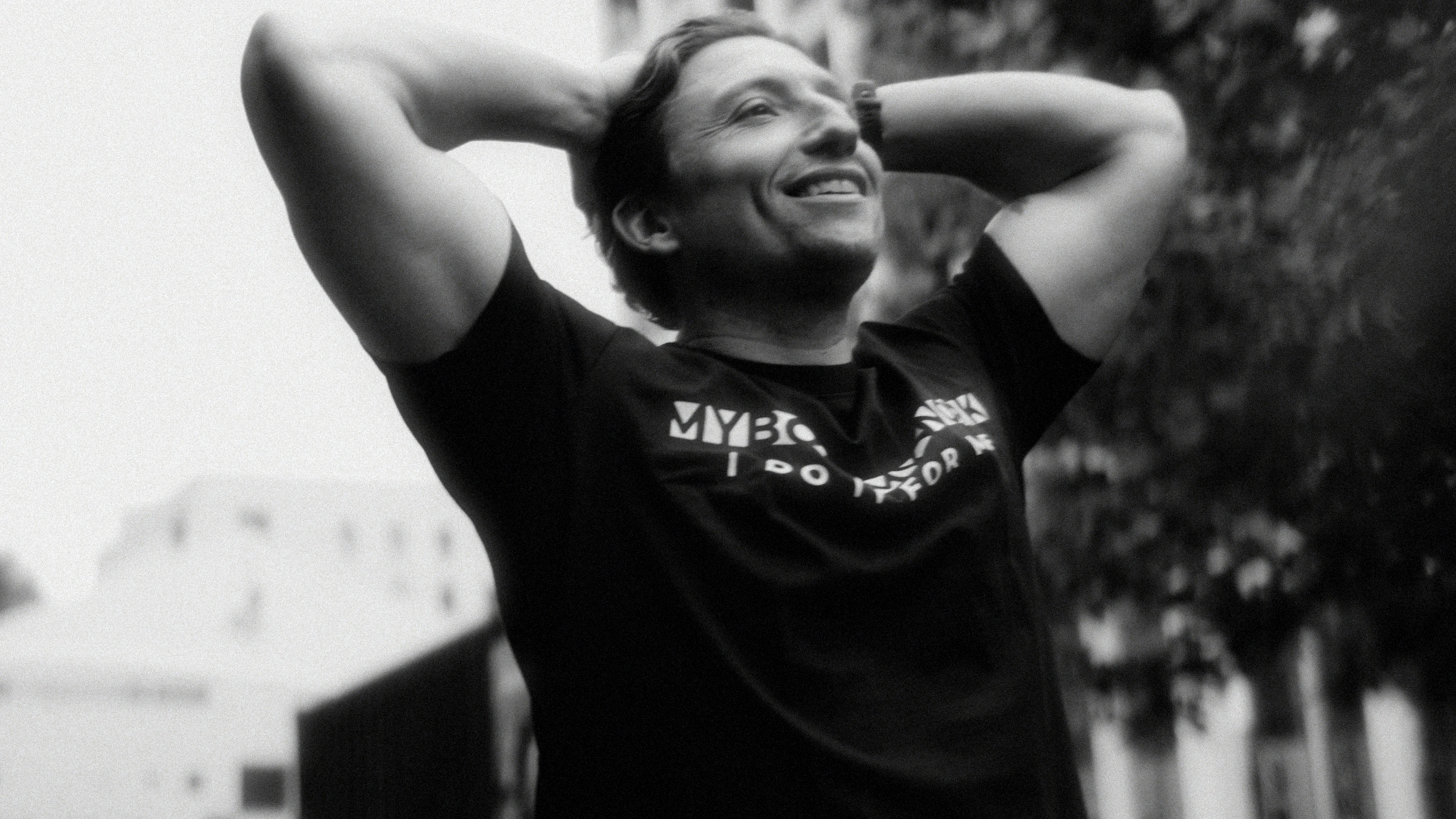 Smiling man with hands behind his head wearing a black shirt outdoors in black and white photo.