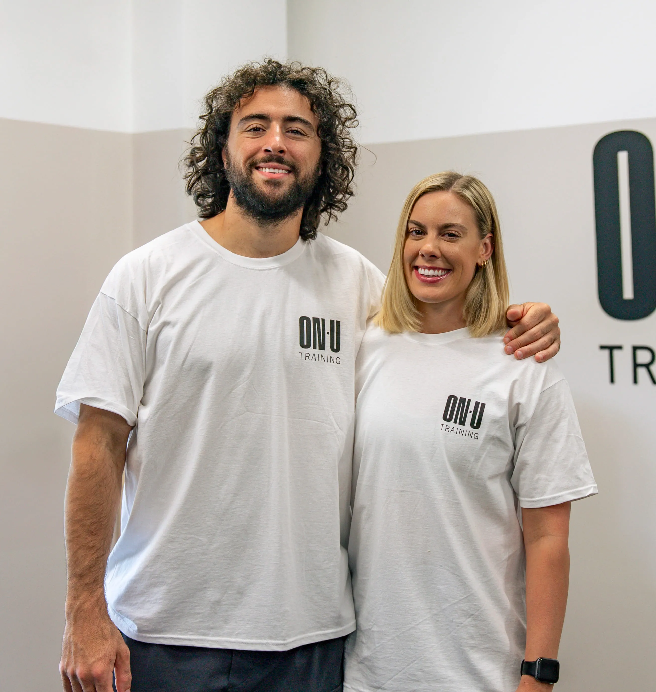 A man and woman smiling, wearing white ON-U Training shirts, standing with arm around each other.