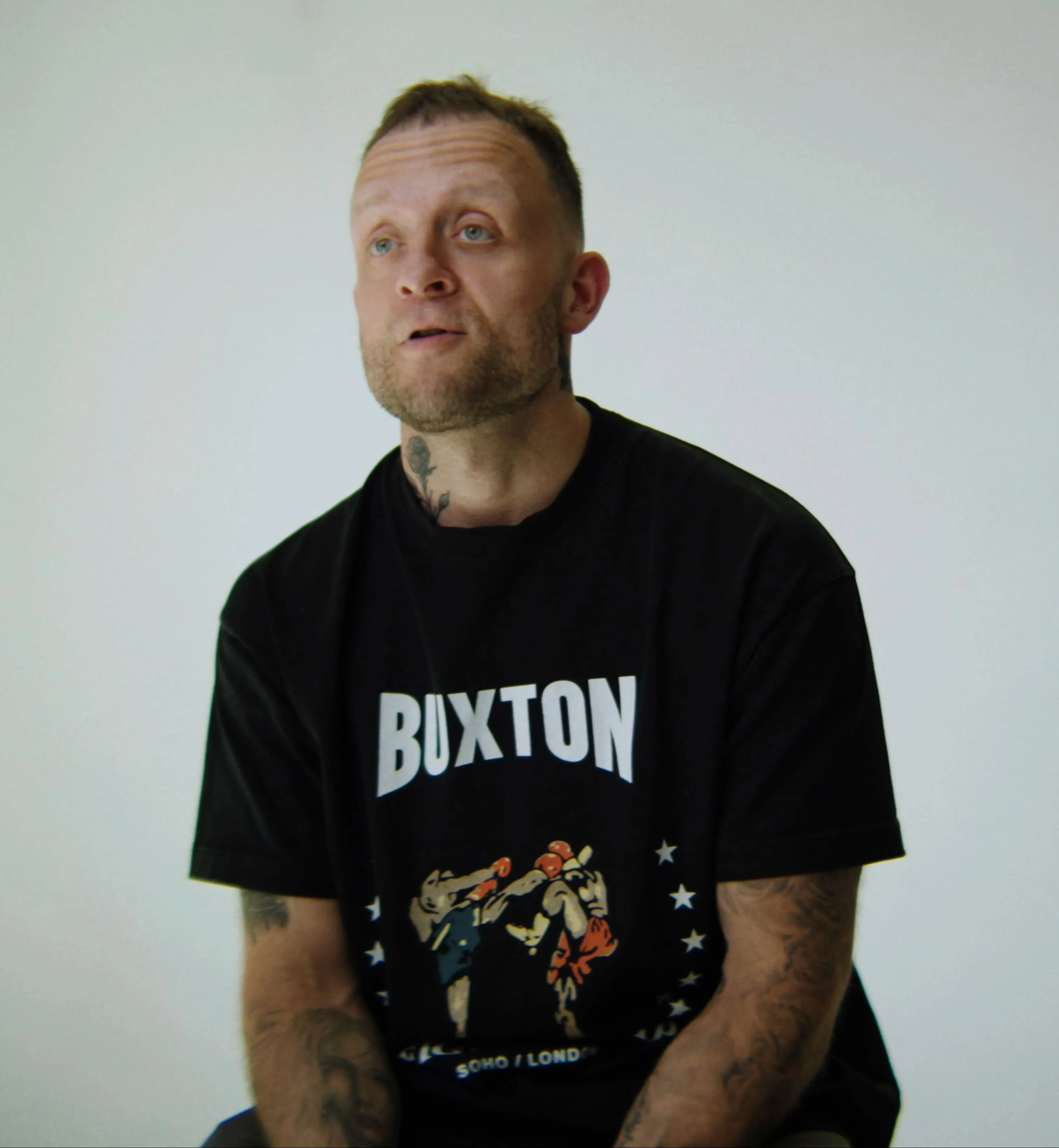 Man with tattoos wearing a black Buxton t-shirt with a boxing graphic on a plain background.