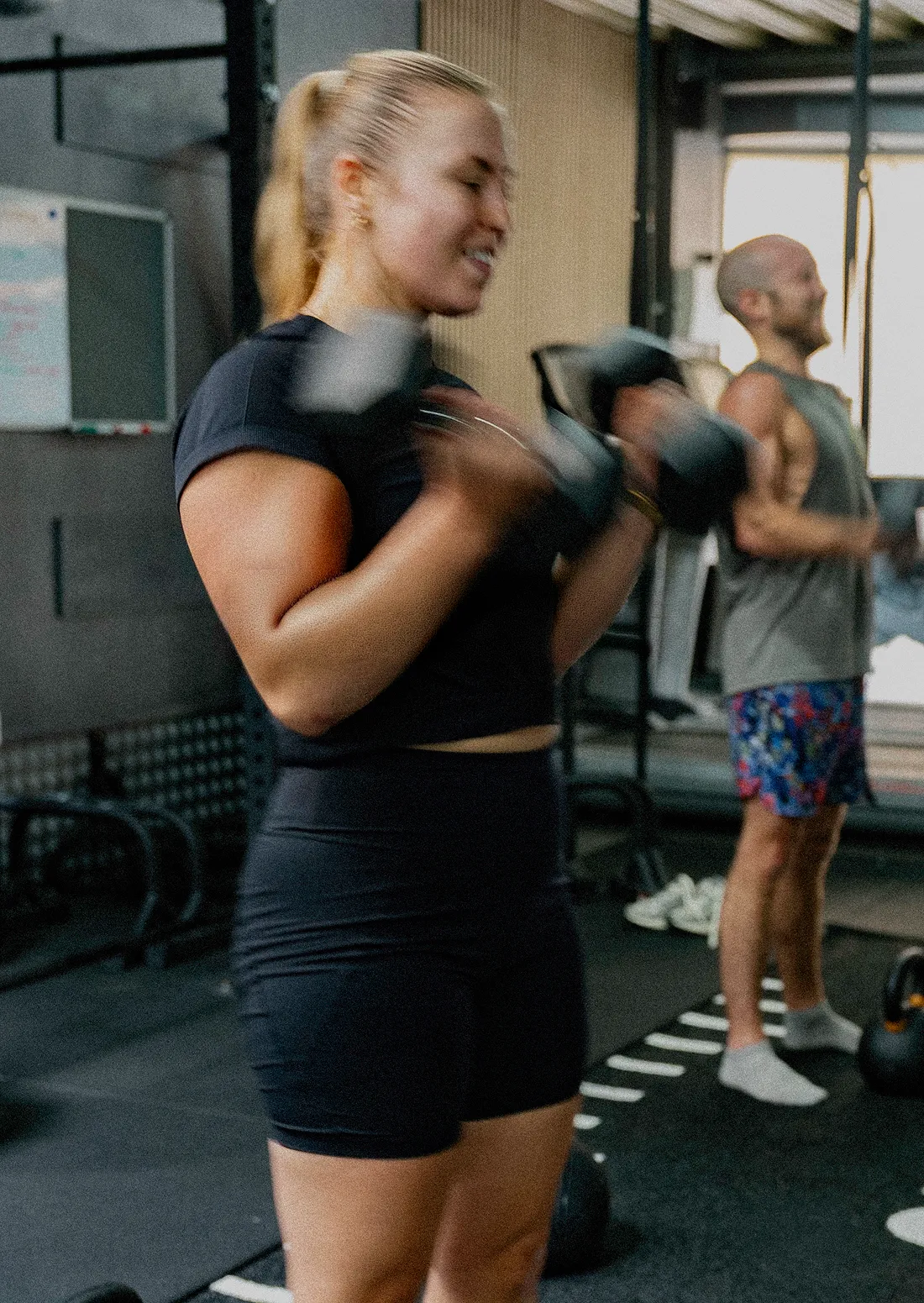 Two people lifting dumbbells in a gym, one woman in black and one man in colorful shorts.