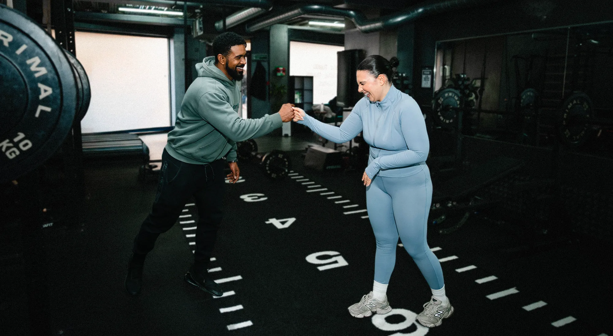 Man and woman in workout clothes fist bumping in a gym with weights and equipment around them.