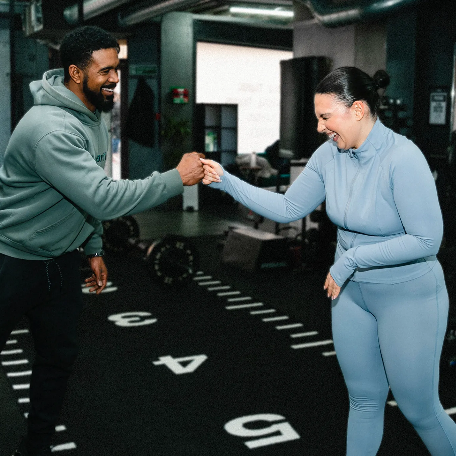 Smiling man and woman fist bumping in gym with exercise equipment in background.