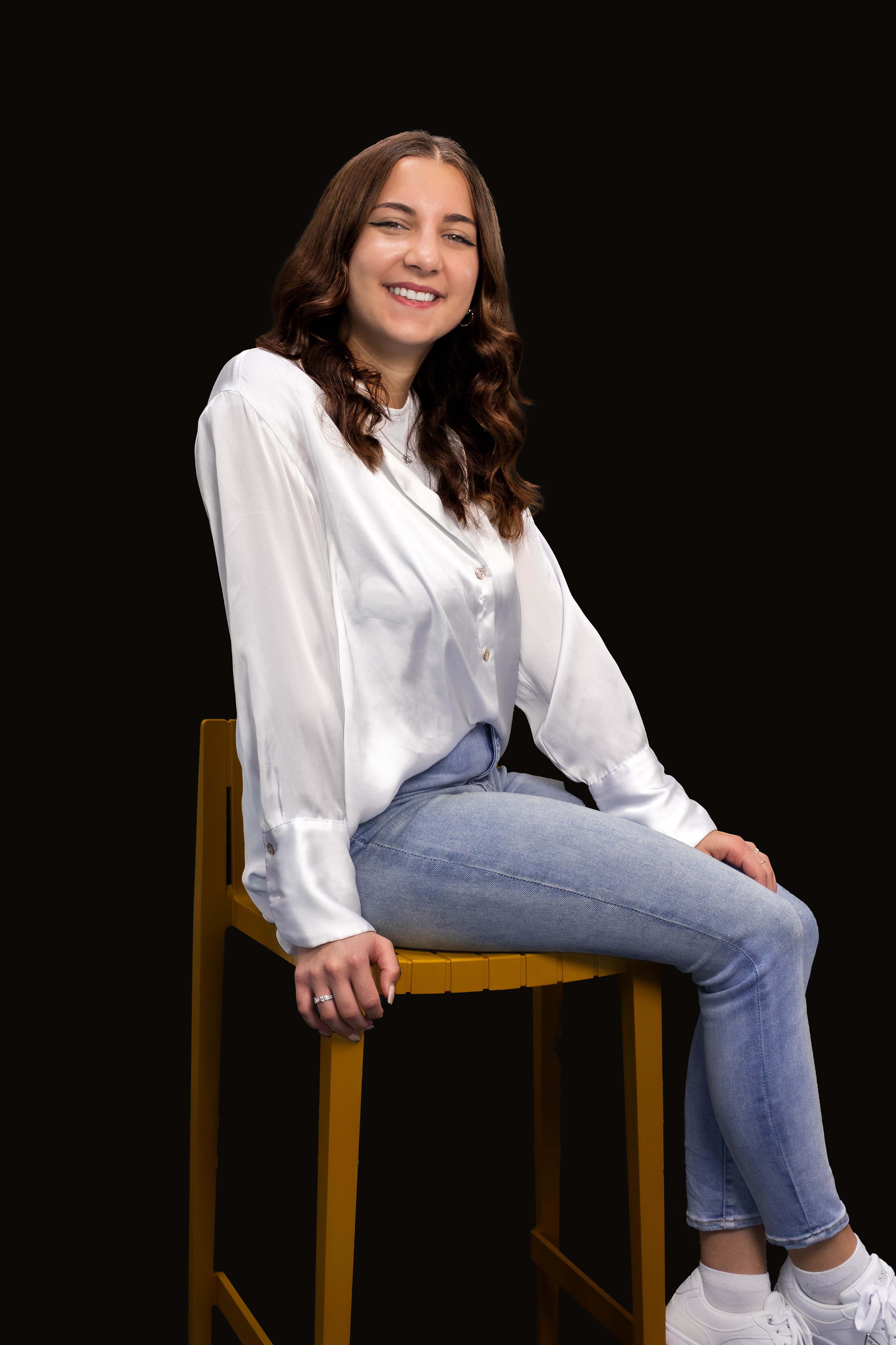 Junge Frau mit langen braunen Locken sitzt auf einem gelben Hocker, trägt weiße Bluse, blaue Jeans und weiße Sneaker, lächelt.