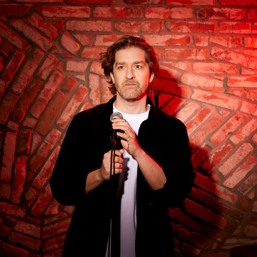 Alex Ploeg met een serieuze blik, een microfoon en een rode muur