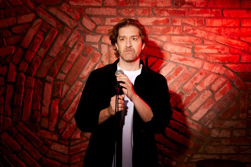 Alex Ploeg met een serieuze blik, een microfoon en een rode muur