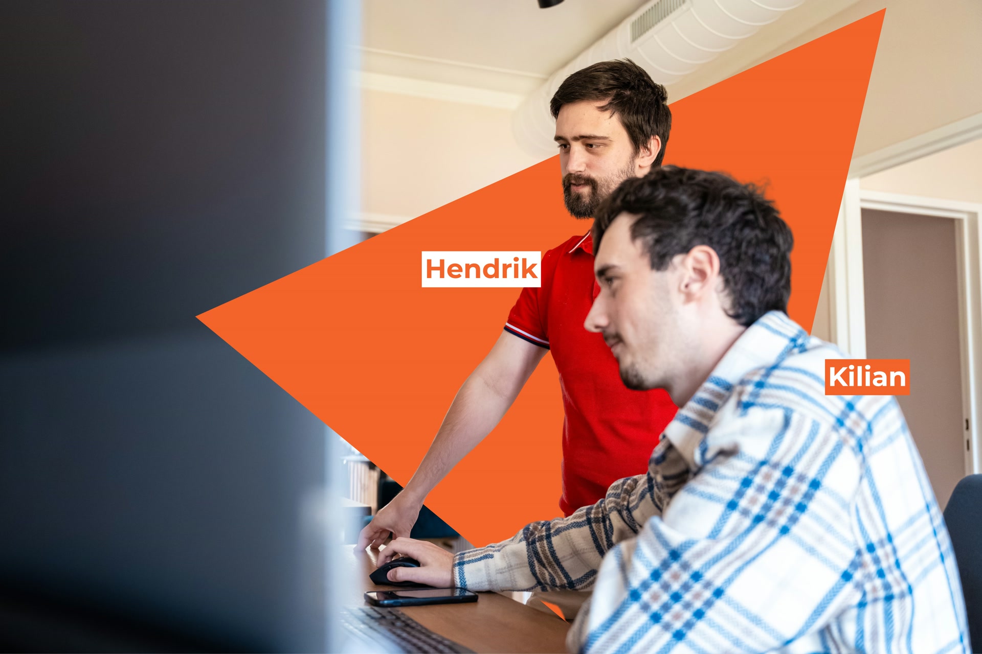 Hendrik geeft uitleg aan Kilian tijdens het werken aan een computerscherm in een moderne werkruimte.
