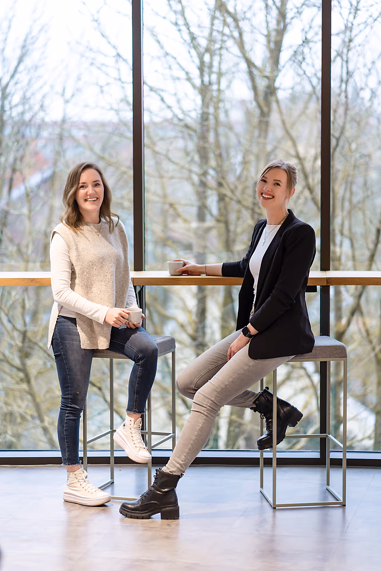 Zwei Personen sitzen an einem Tisch, halten Tassen in der Hand und lächeln in die Kamera