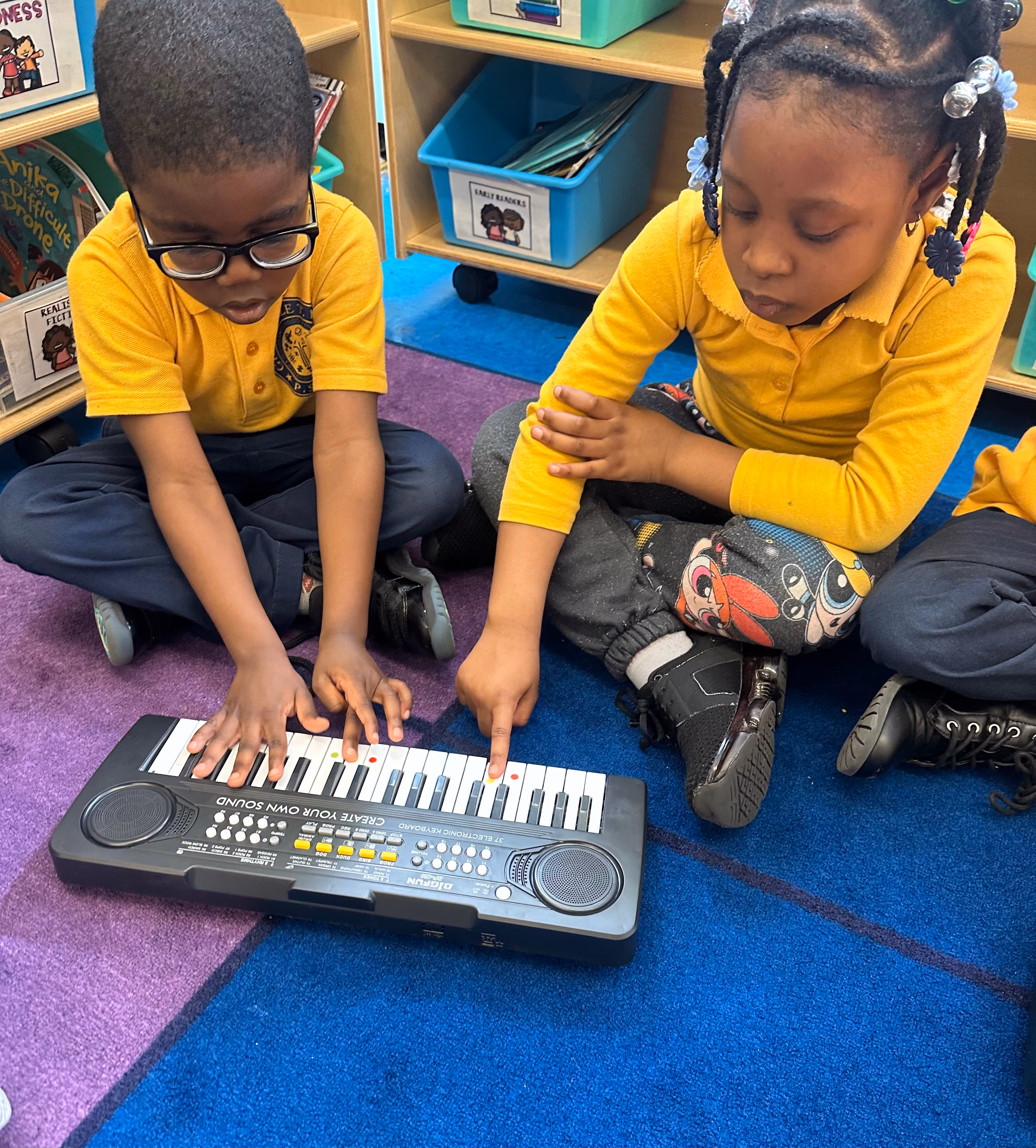 Two young students in uniform playing a small keyboard together on classroom floor