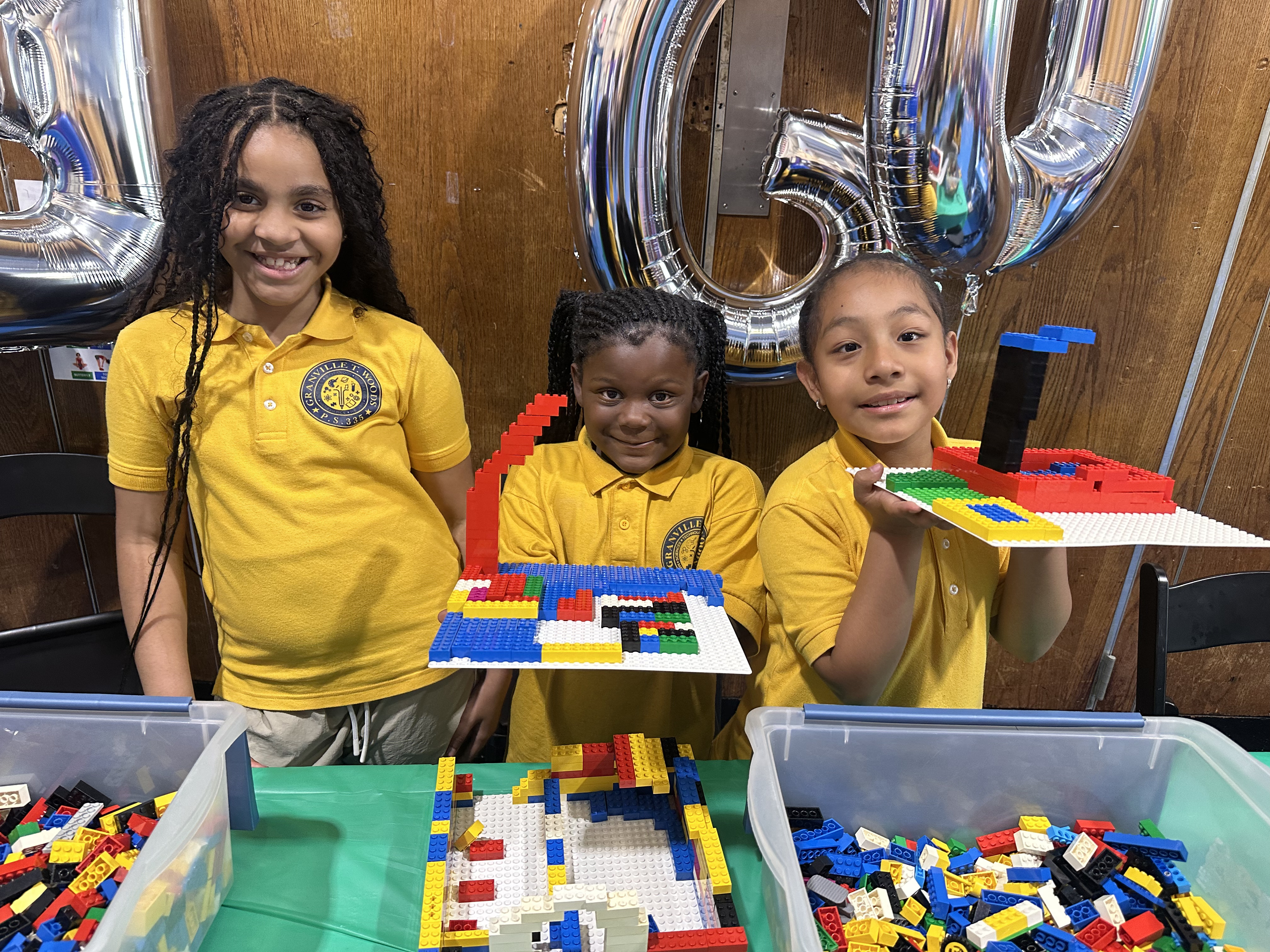 Three students in yellow uniforms proudly displaying colorful LEGO creations