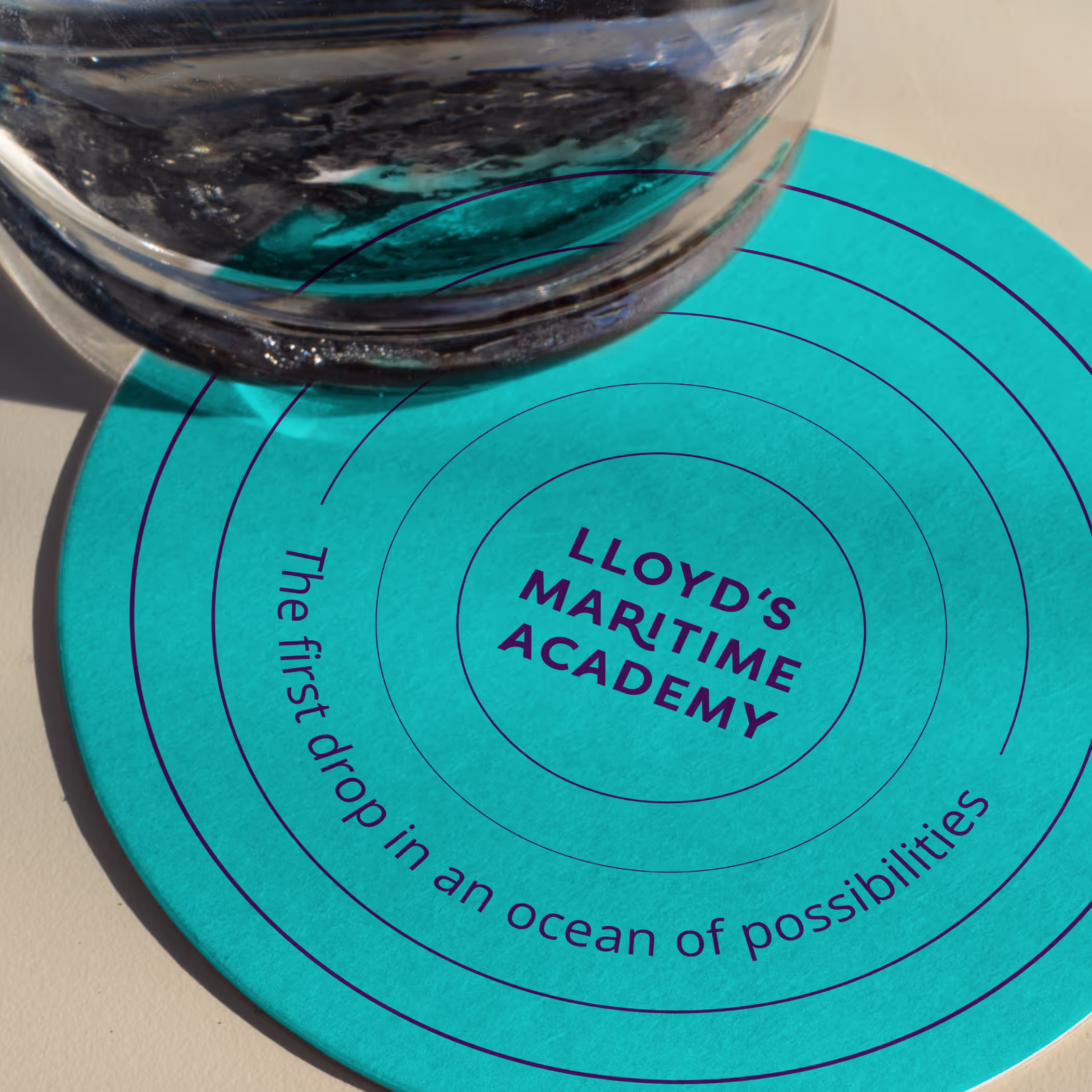 A glass on a coaster with LMA branding and the words "the first drop in an ocean of possibilities"