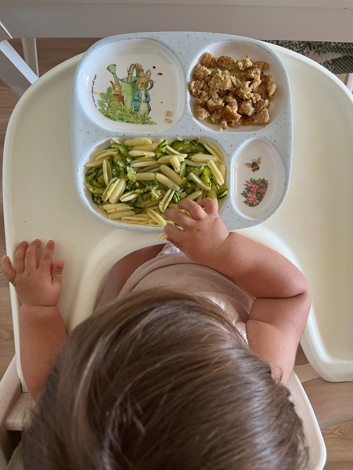 Bambino seduto su un seggiolone che mangia pasta corta con zucchine da un piatto diviso decorato con coniglietti e fiori.
