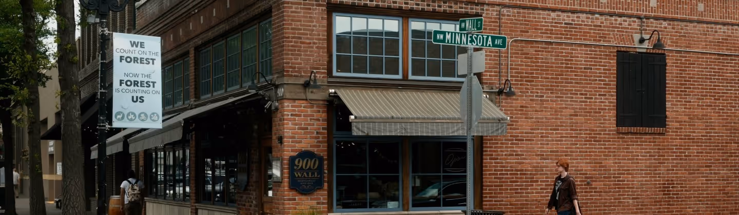 Brick building on the corner of NW Minnesota Avenue and Wall Street with a sign reading '900 Wall Restaurant & Pub' and a banner about forest conservation, two people walking nearby.