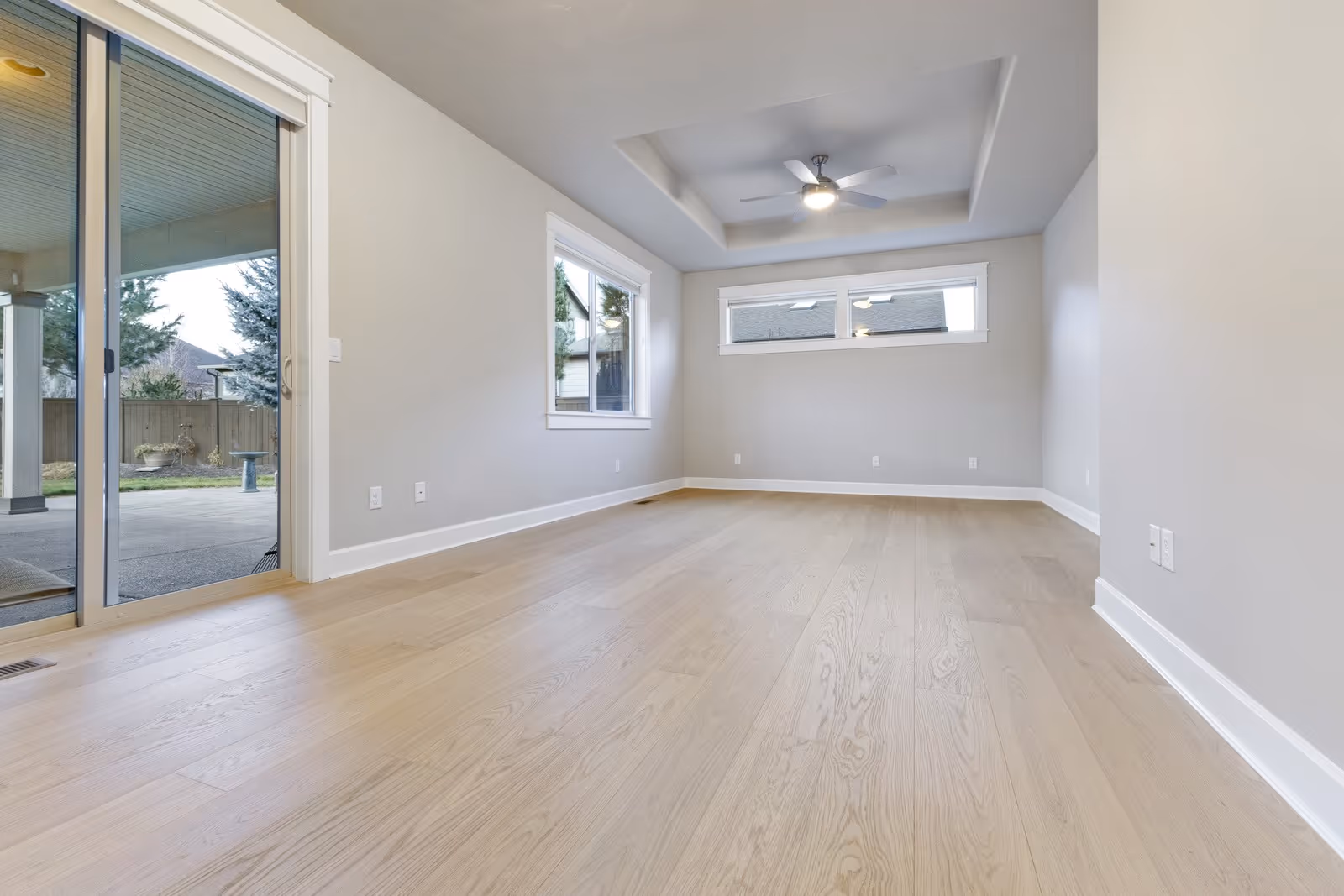 Empty modern room with light wood flooring, gray walls, ceiling fan, rectangular and square windows, and sliding glass door leading to a backyard patio.