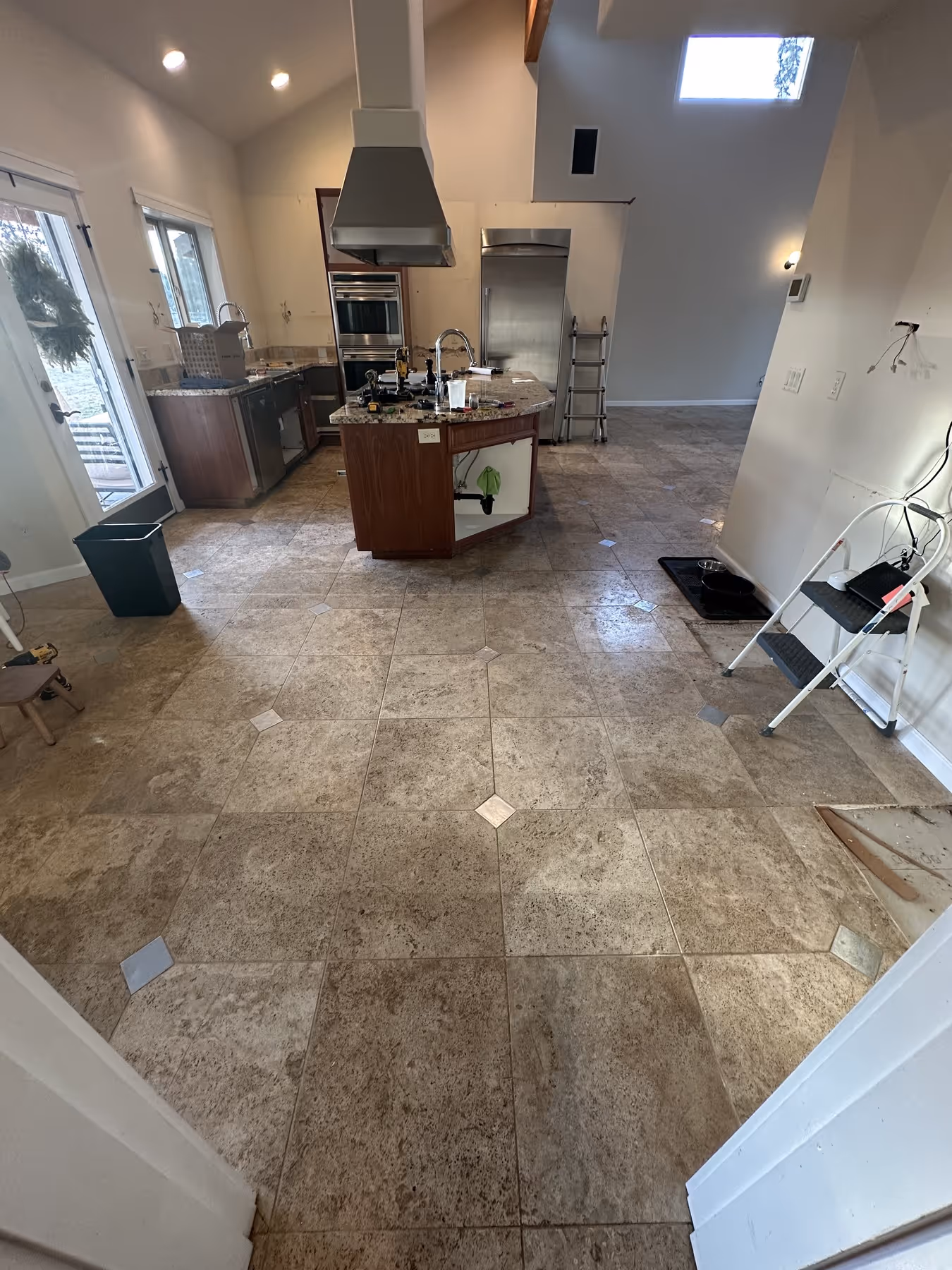 Kitchen under renovation with tiled floor, central island with open cabinet showing plumbing, stainless steel appliances, step ladder, and tools on the island.