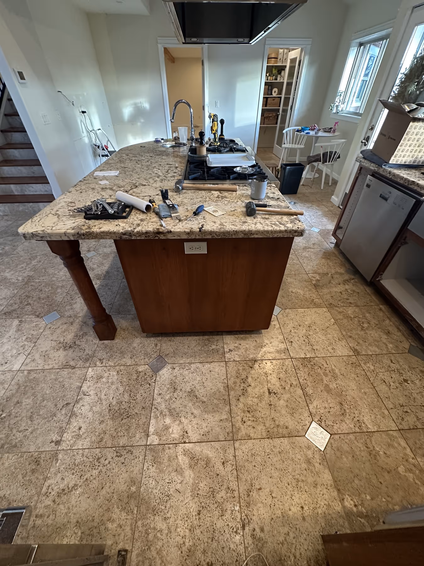 Kitchen island with granite countertop covered in tools, including hammers, screwdrivers, and a paint roller, in a home under renovation. Rustic tile flooring.