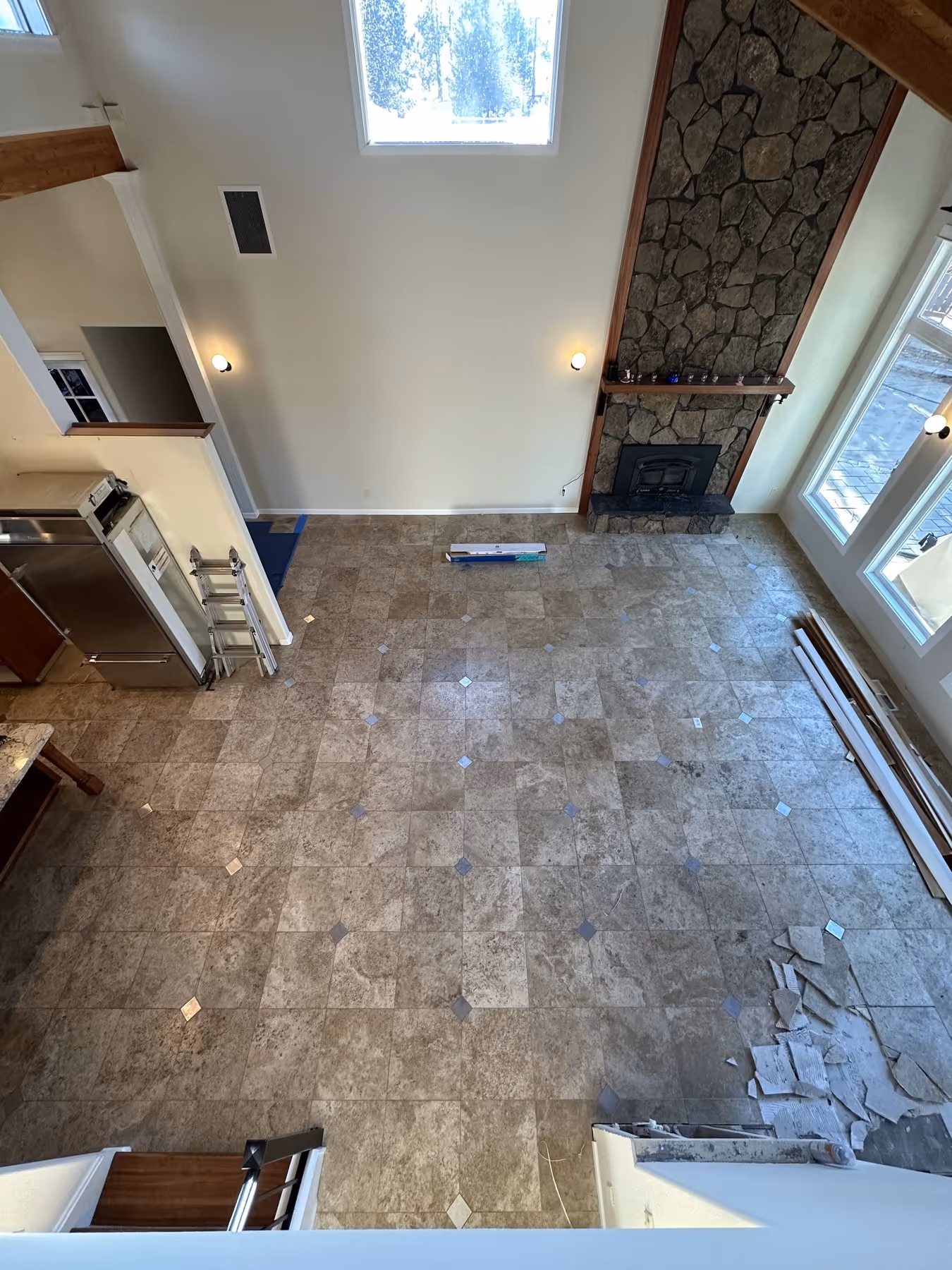 View from above of a tiled floor in a spacious room with stone fireplace, large windows, and scattered construction materials.