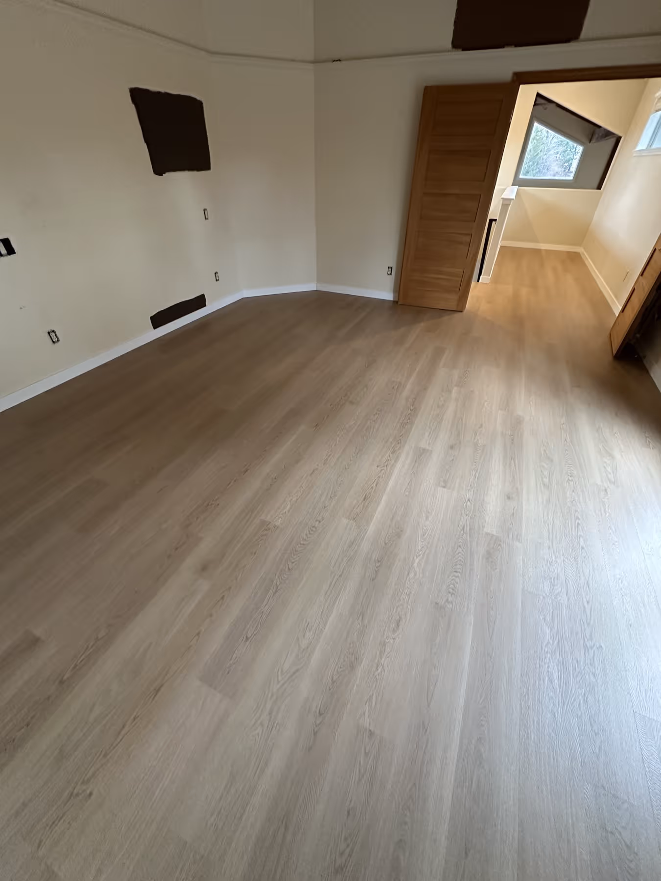 Empty room with newly installed light LVP flooring, beige walls, and an open wooden door leading to a nearby space with a window and similar flooring.