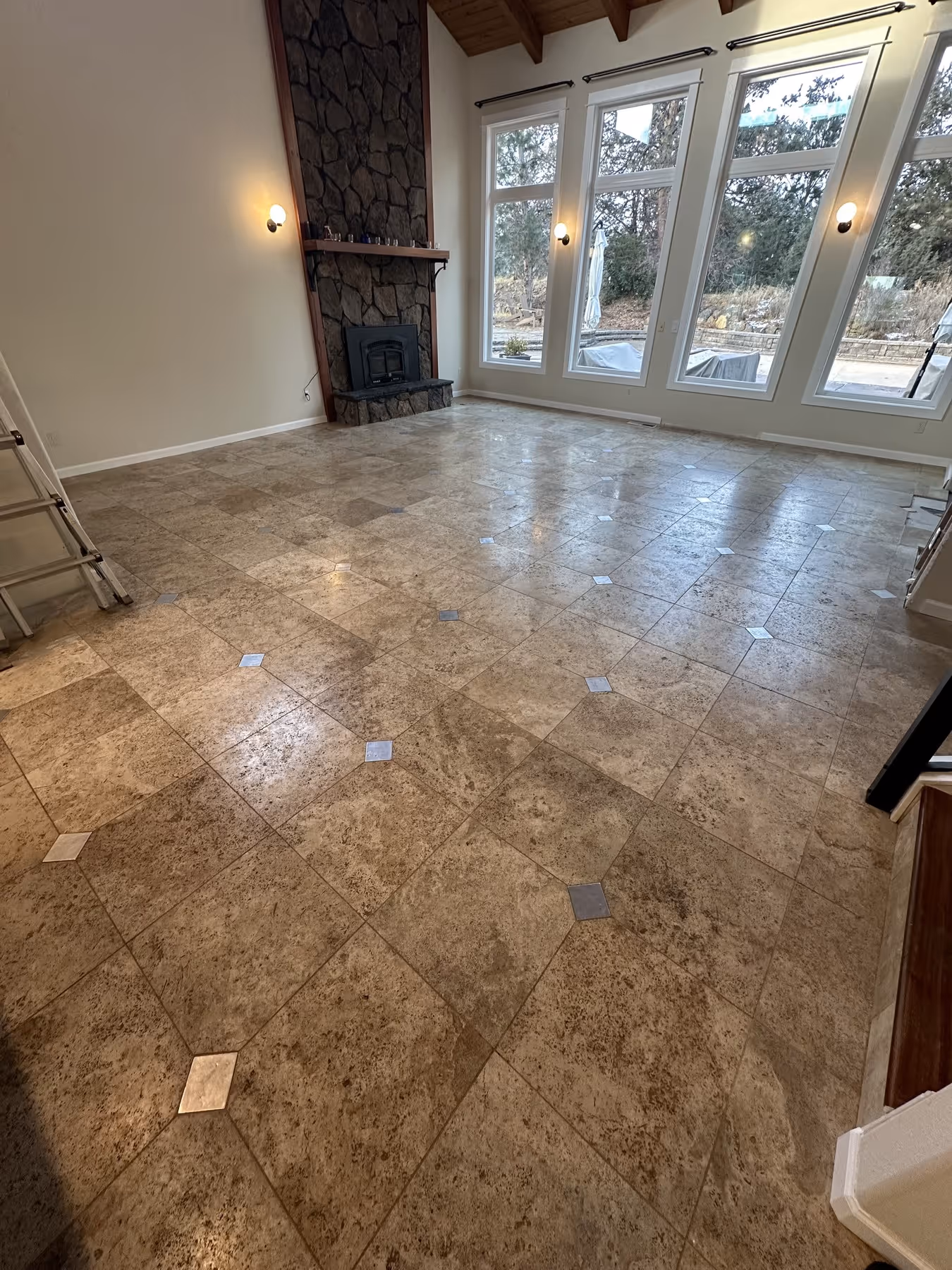 Spacious room with stone fireplace, beige tiled floor with small square accents, and large windows overlooking an outdoor patio.