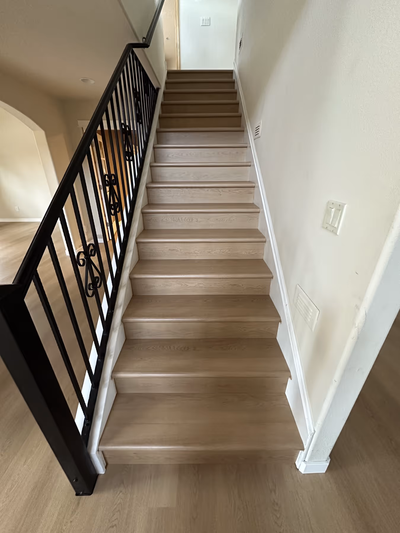 Light LVP flooring staircase with black metal railing and decorative scrolls on the left, and white walls on the right.