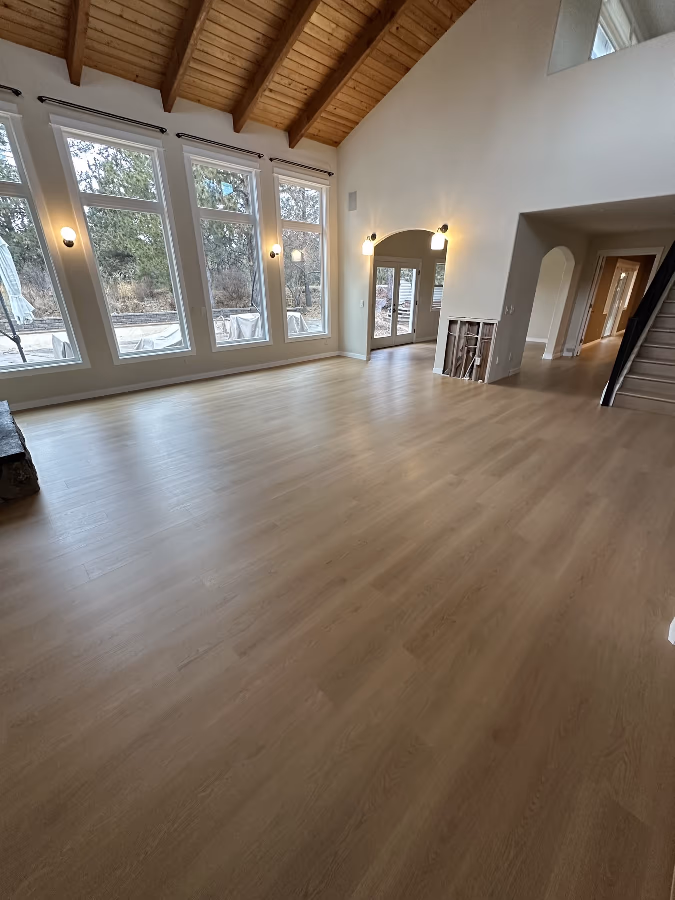 Spacious empty room with high wooden ceiling, large windows, light LVP floor, and a stairway on the right.