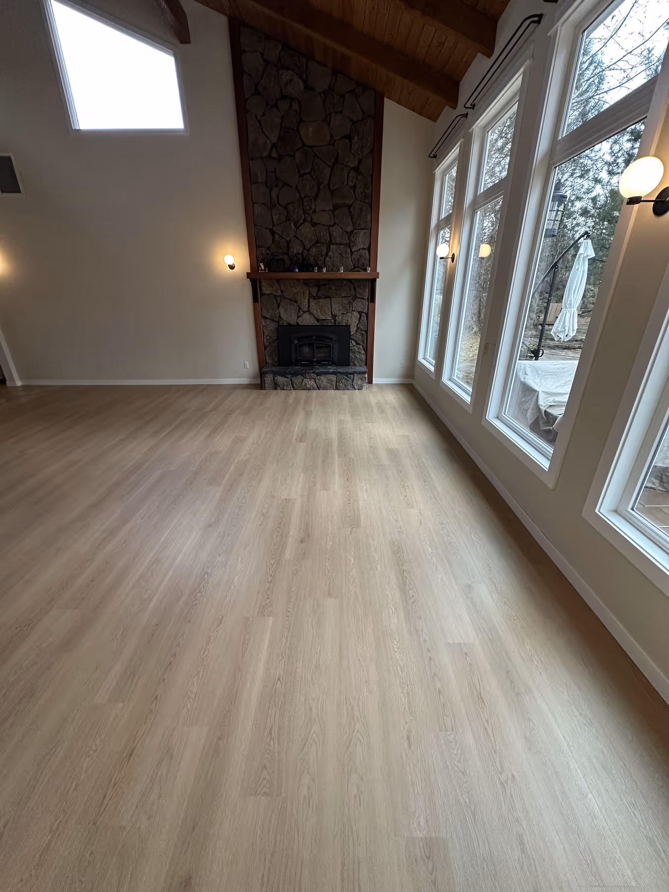 Empty living room with light LVP flooring, stone fireplace, large windows, and wood-paneled vaulted ceiling.