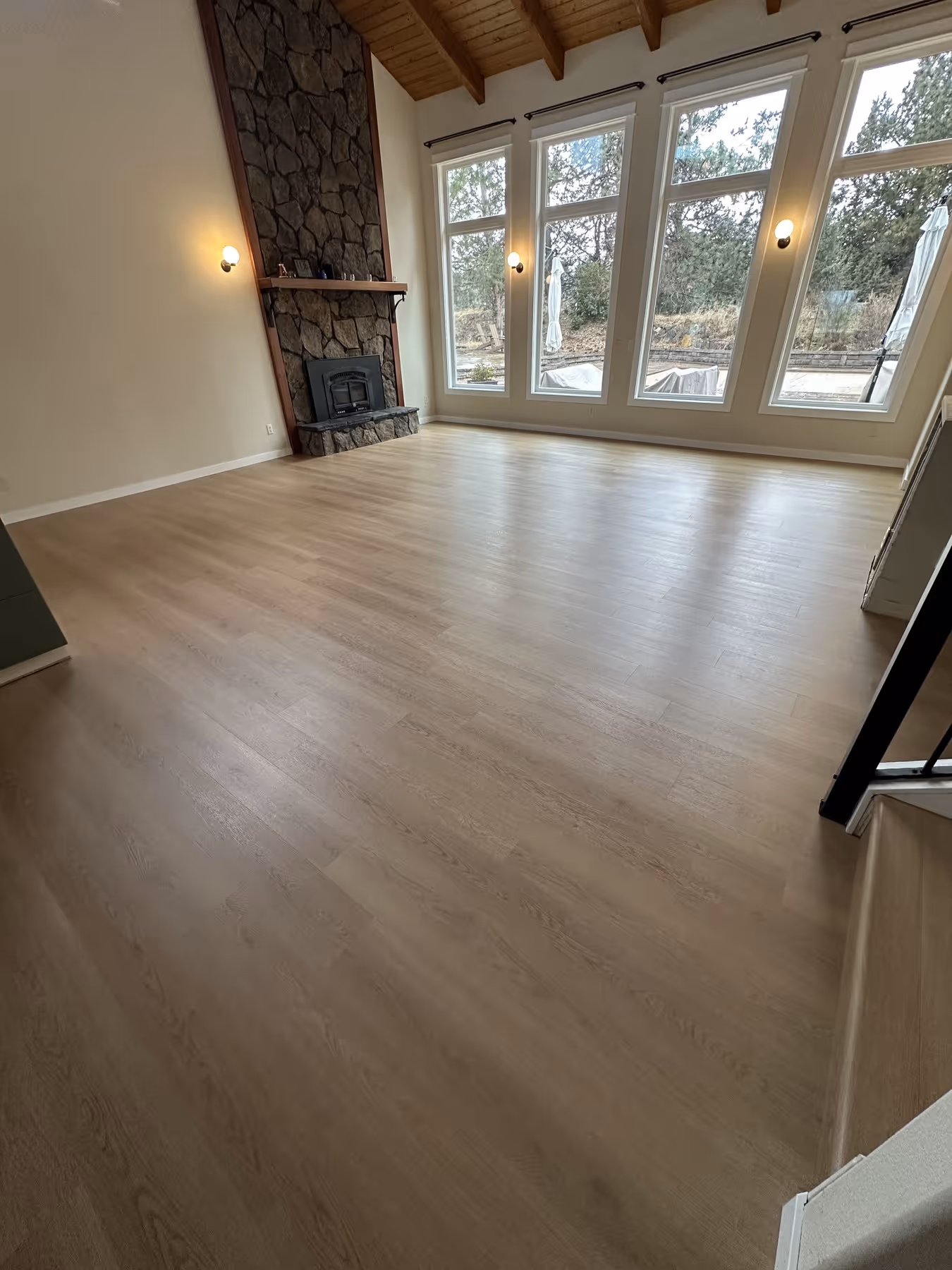 Empty room with light LVP flooring, stone fireplace, and large windows overlooking an outdoor area.
