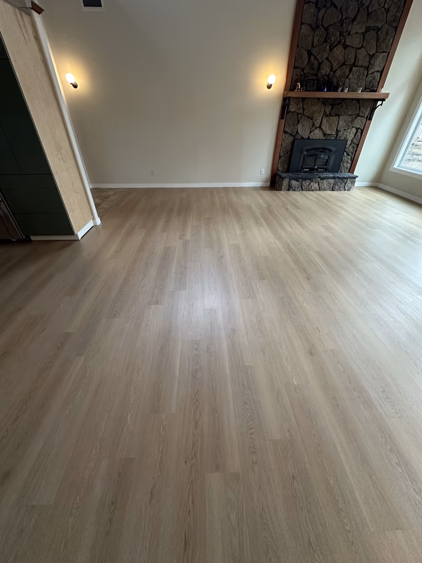 Empty room with light LVP flooring, stone fireplace, and large window.