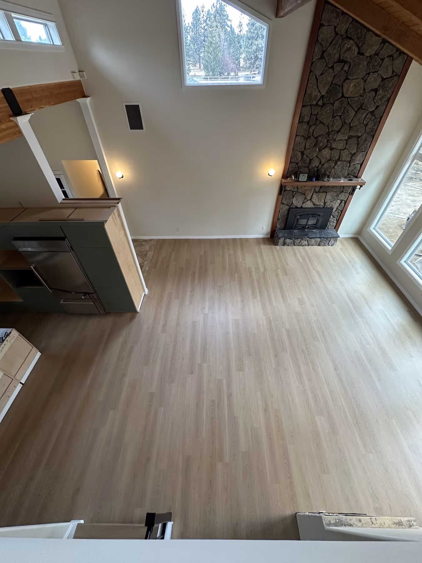 Empty living room with light LVP flooring, a stone fireplace, high ceiling, and large windows.