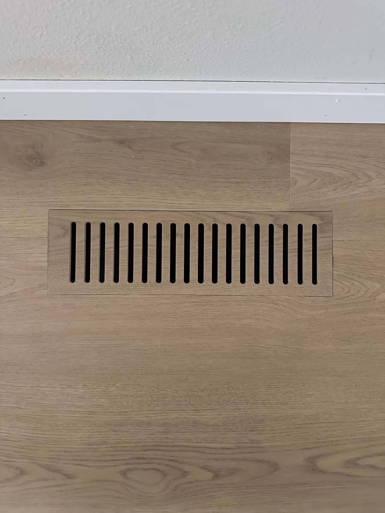 Wooden floor vent with vertical slats embedded in light wood flooring beneath a white baseboard.