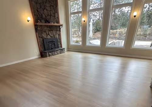 Empty room with light LVP flooring, stone fireplace on the left, three large windows, and wall sconces.