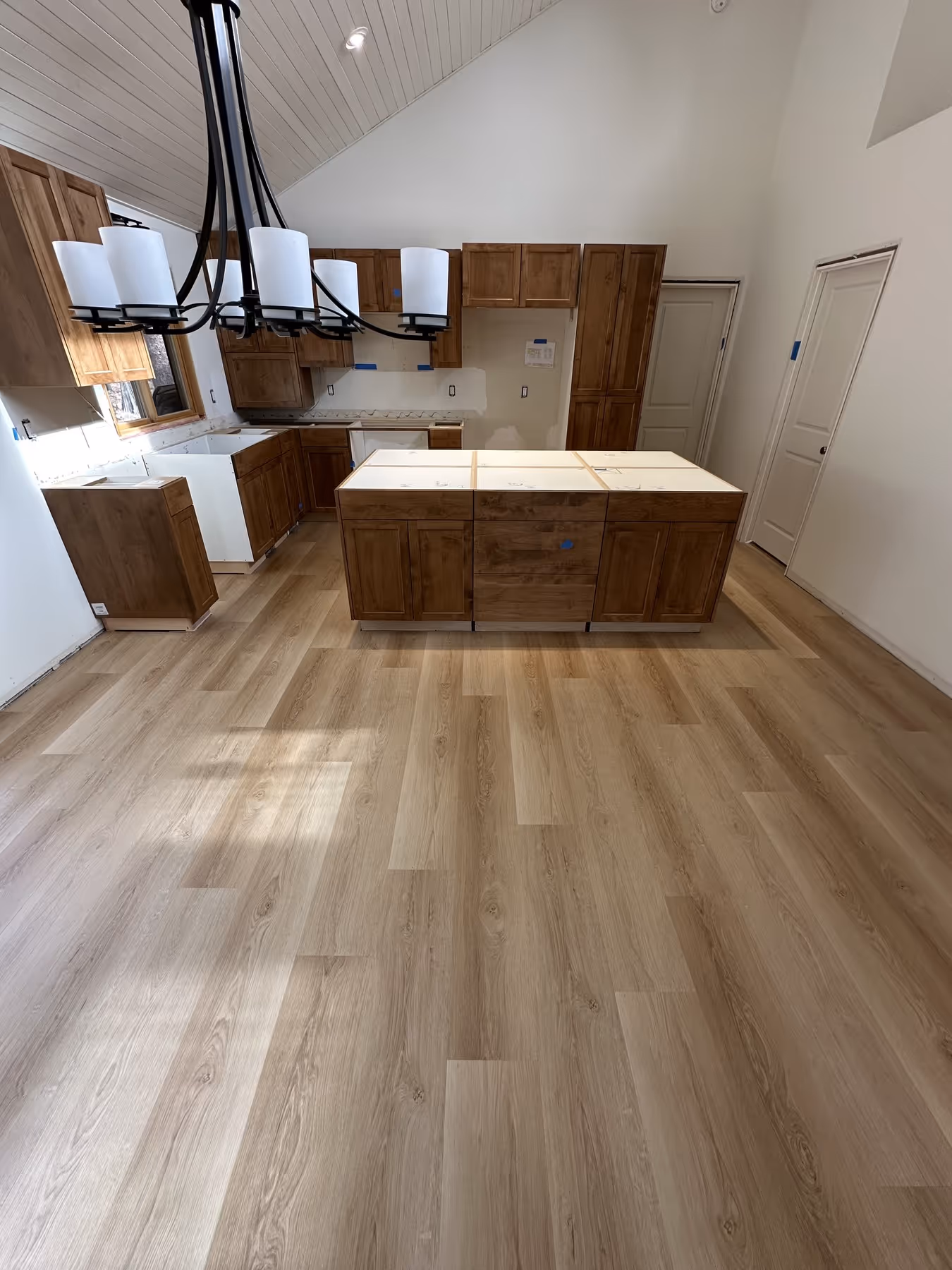 Unfinished kitchen with newly installed wooden cabinets, island, laminate flooring, and a modern black chandelier with six white shades.