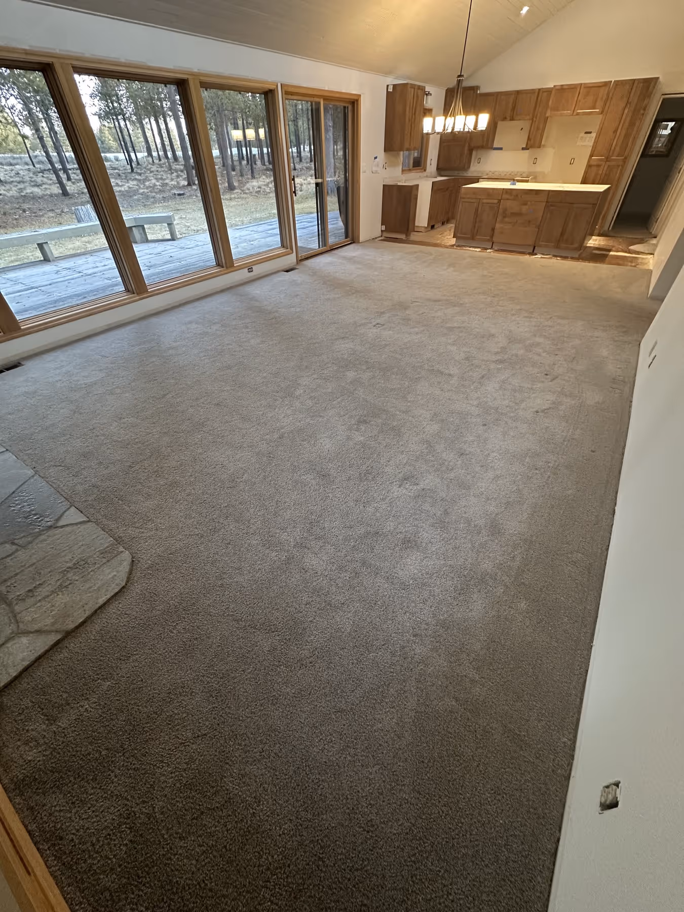 Empty living room with beige carpet, large windows and sliding door looking out to a wooded backyard, and an adjacent kitchen with wooden cabinets and a hanging light fixture.