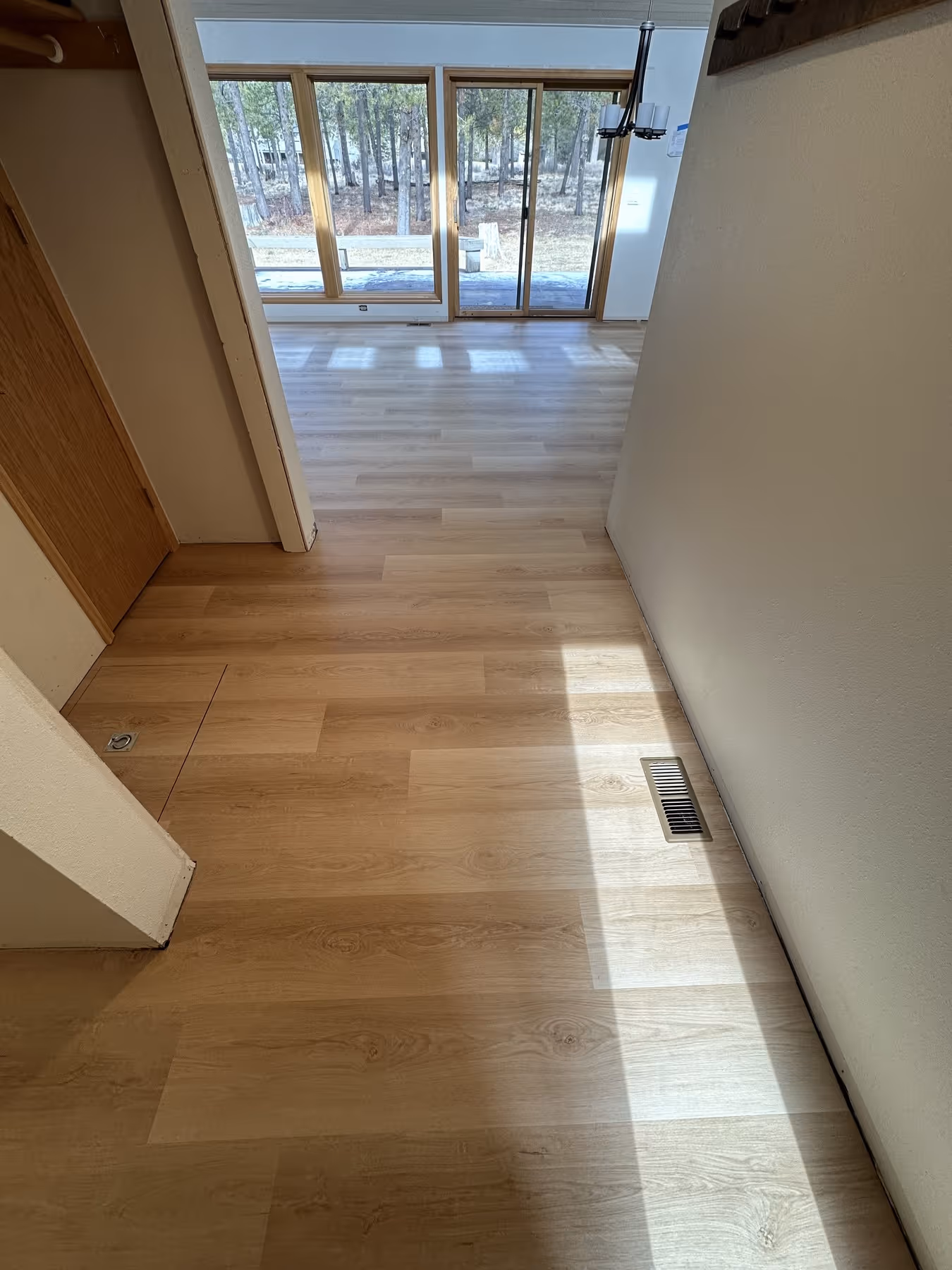Indoor space with light wood flooring, white walls, floor vent, and large windows with sliding glass door showing a wooded outdoor area.