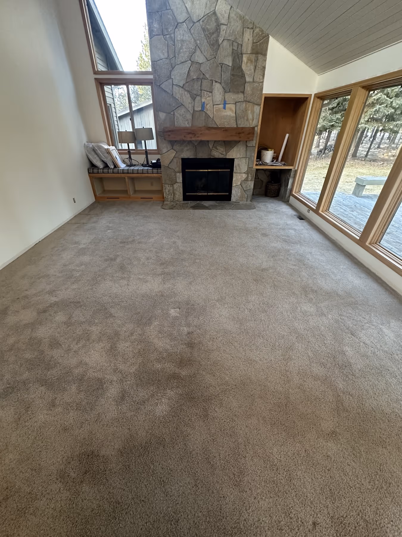 Living room with beige carpet, large stone fireplace with wooden mantel, built-in bench with cushions, and large windows looking out to a wooded yard.