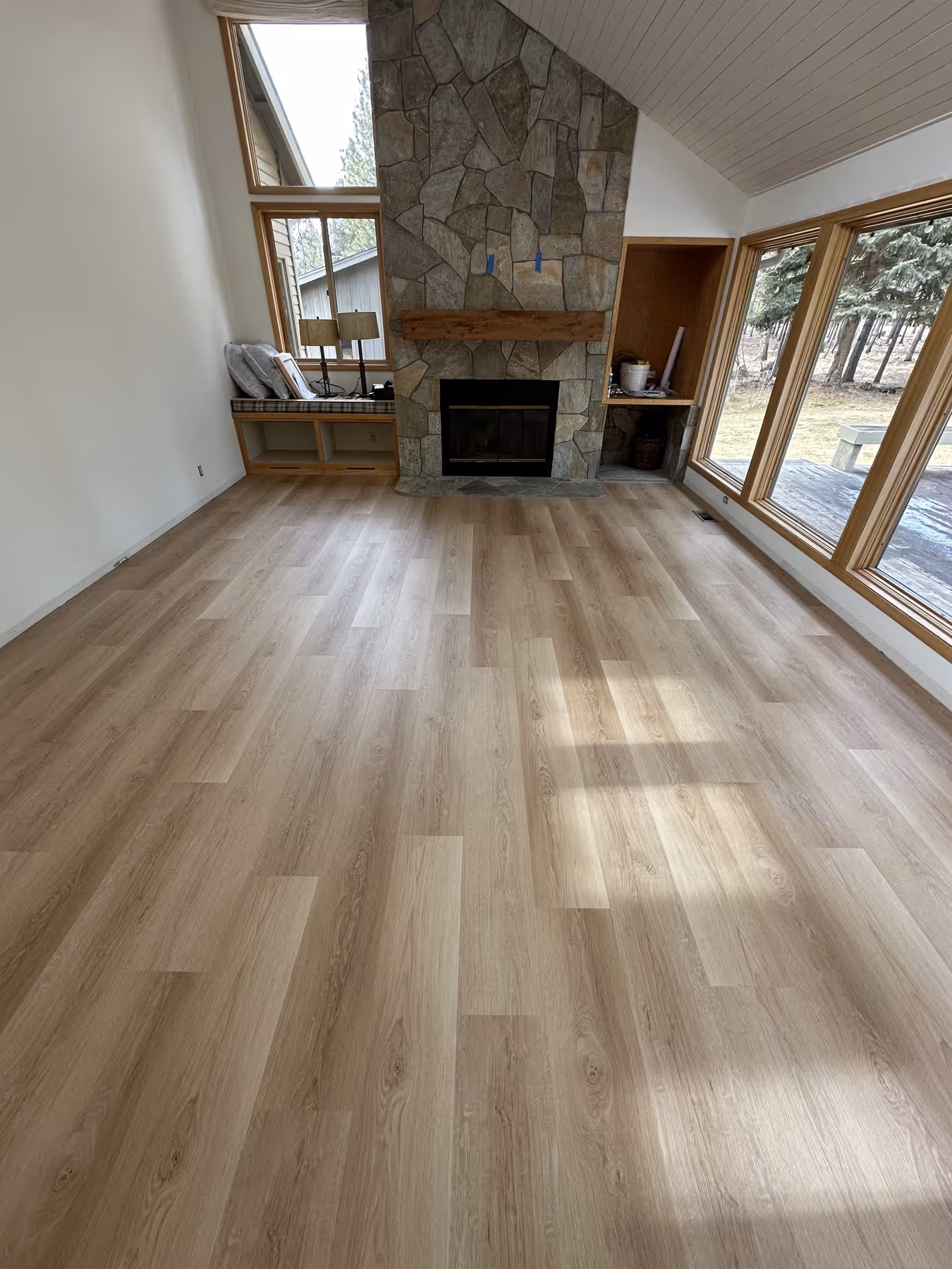 Empty room with light wood flooring, large stone fireplace, built-in shelves, and windows overlooking a wooded yard.