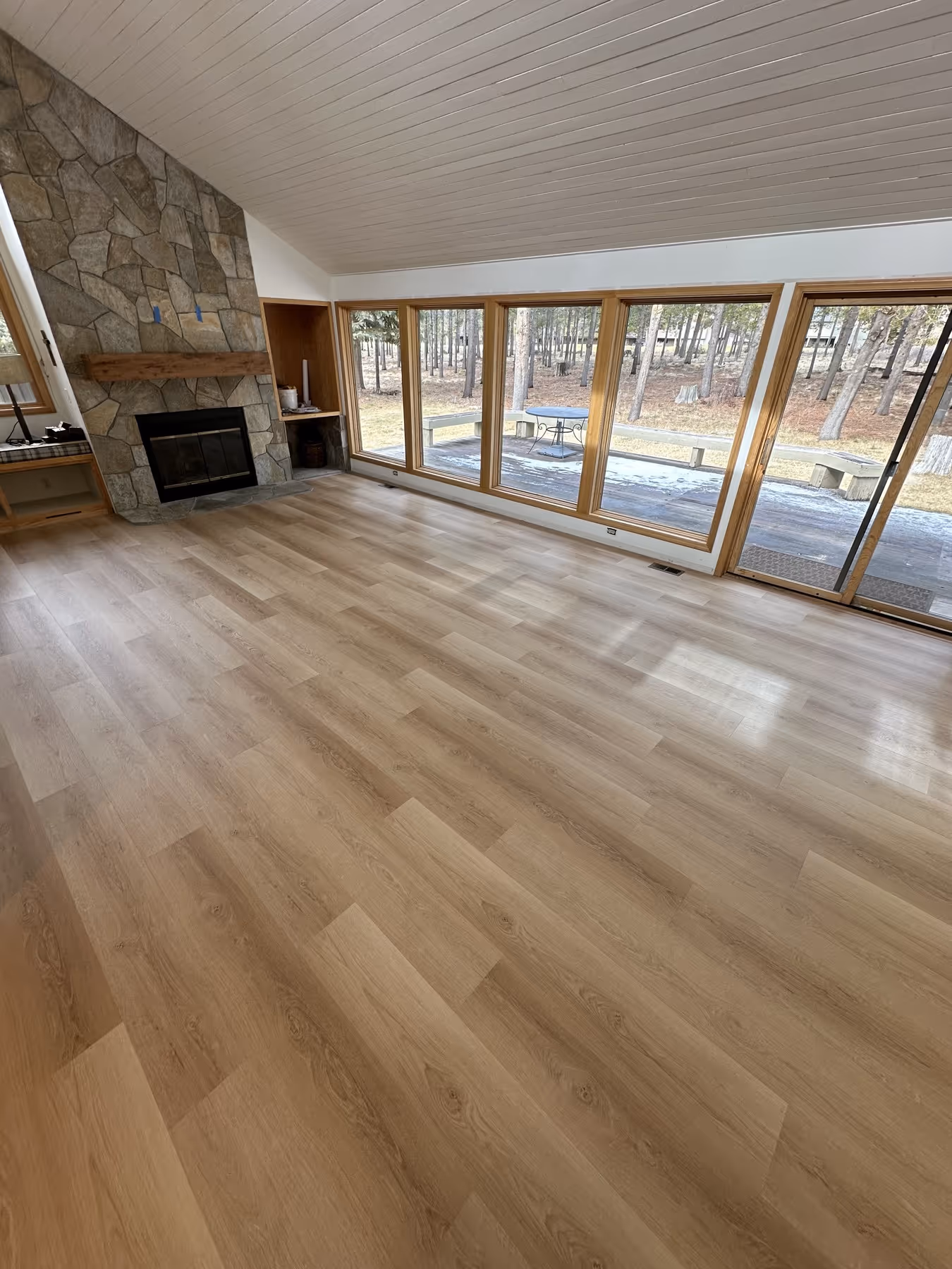 Empty room with light wood flooring, stone fireplace, wood paneled ceiling, and large windows looking out to a wooded yard.