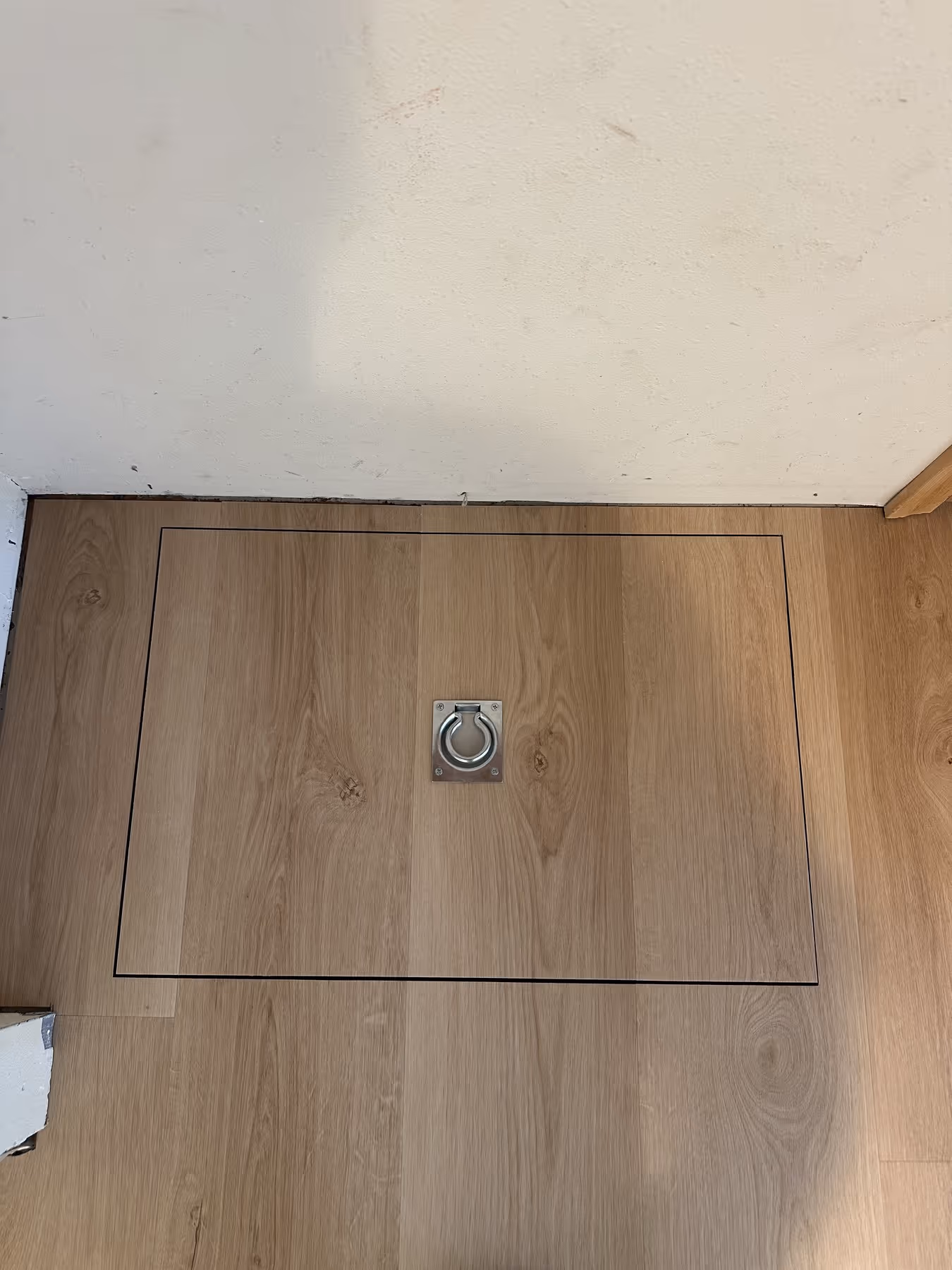Wooden floor panel with a square hatch outlined in black and a metal ring handle in the center.