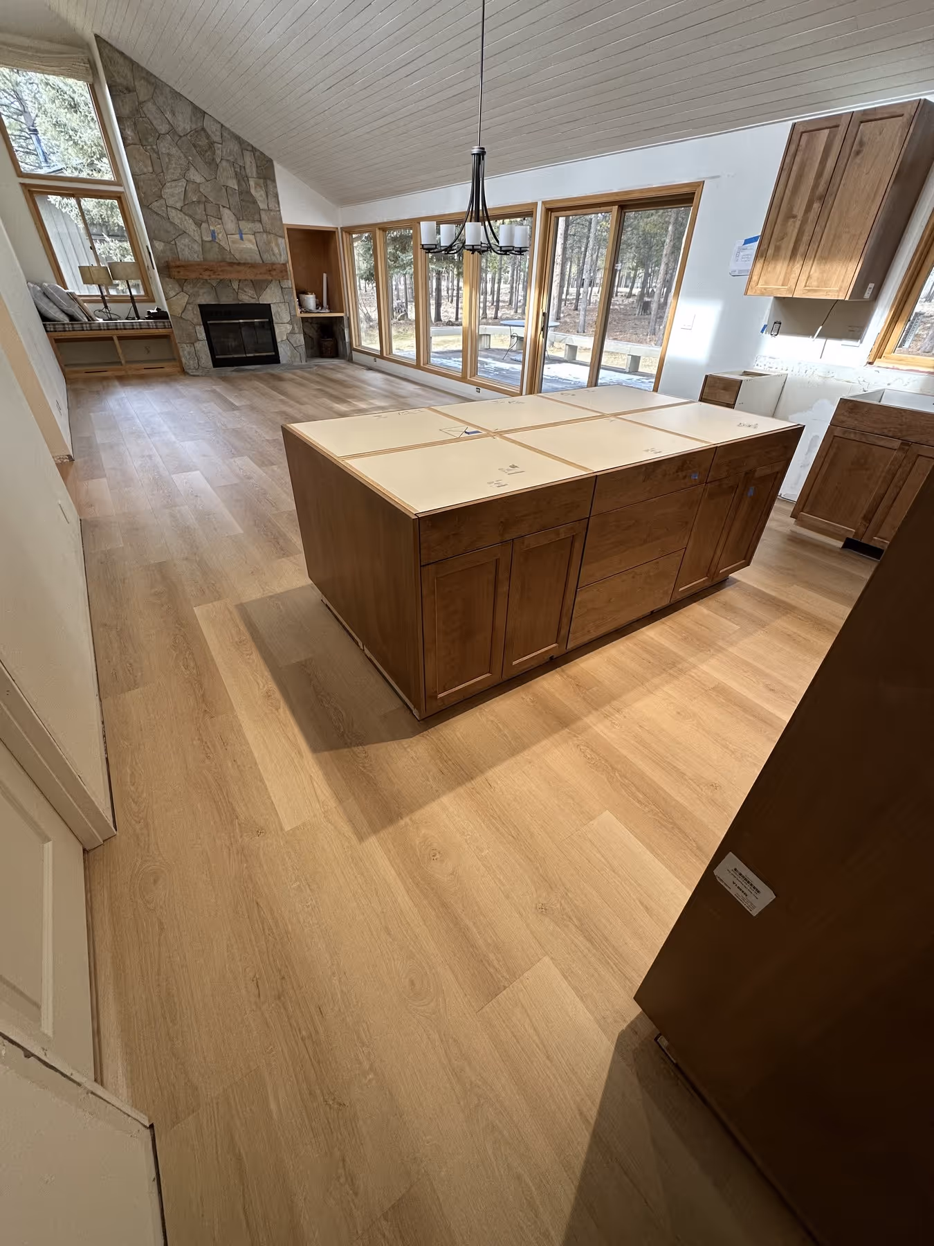 Open kitchen and living room with light wood flooring, a wooden kitchen island without a countertop, stone fireplace, large windows, and a chandelier.