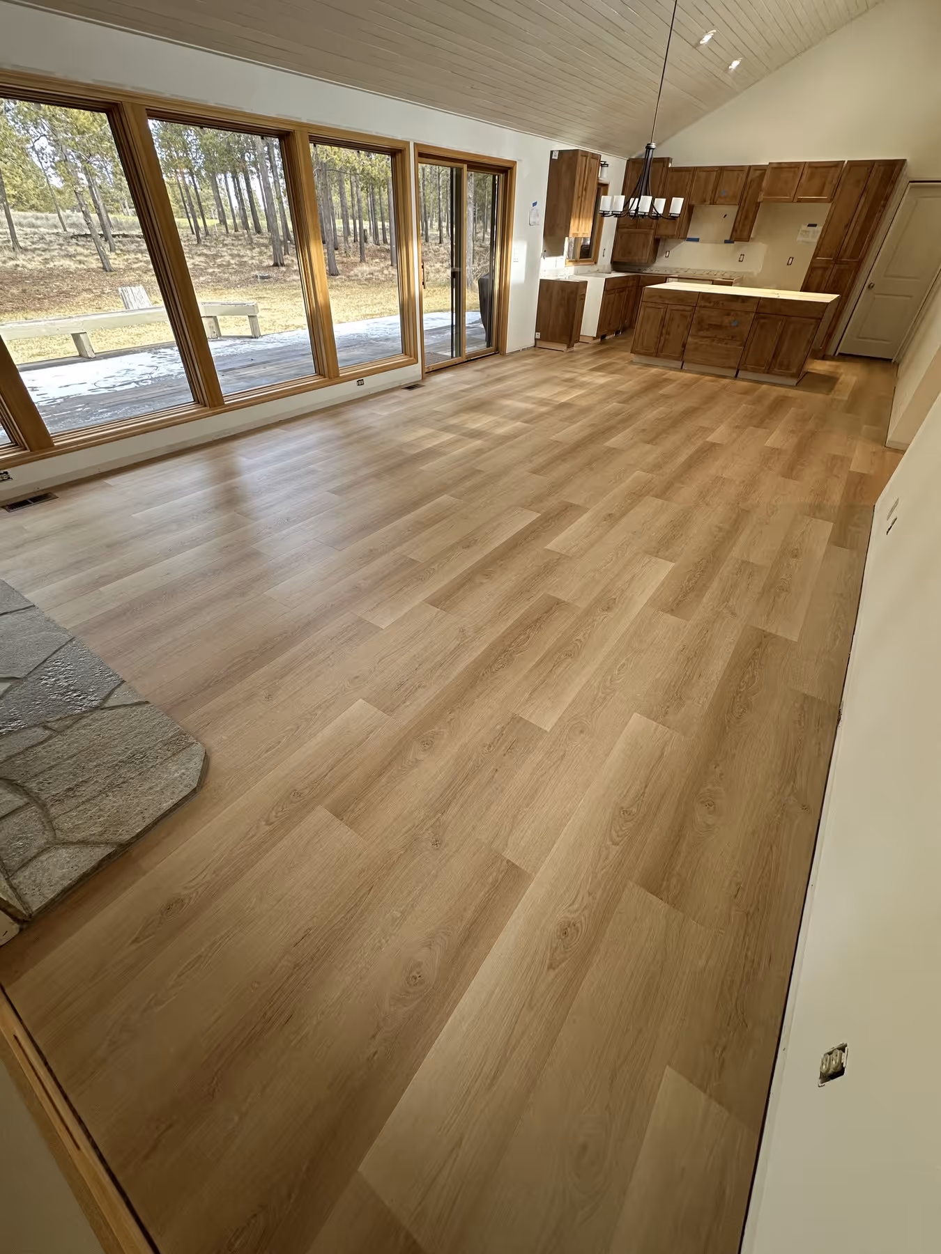Open room with light wood laminate flooring, large windows overlooking trees, and a kitchen with wooden cabinets and island.