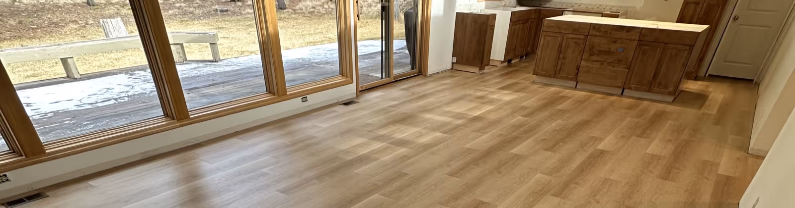 Empty room with large floor-to-ceiling windows, light wood flooring, and unfinished wooden cabinets along one wall.