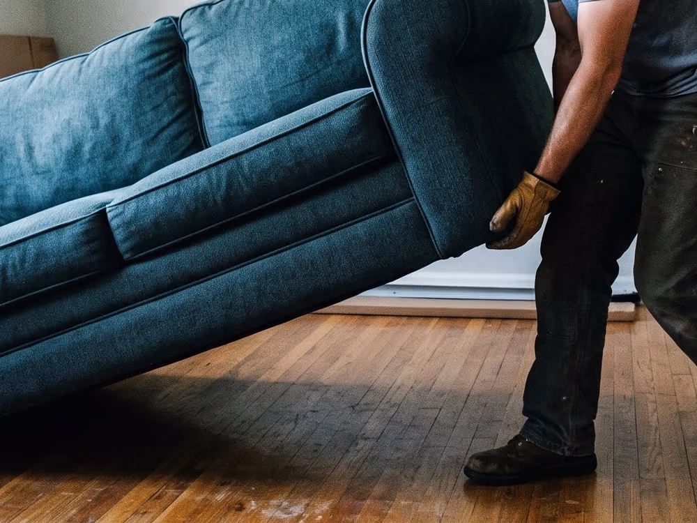 Person wearing gloves lifting a dark blue sofa on a wooden floor indoors.