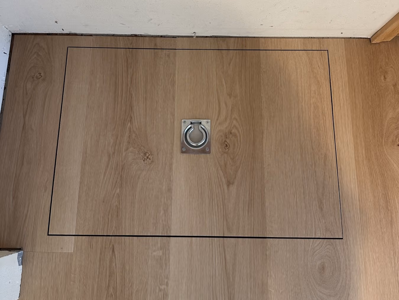 Wood floor panel with a metal ring pull handle set inside a rectangular outline.