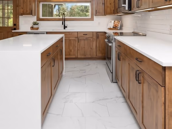 Modern kitchen with wooden cabinets, white countertops, a large island, marble-style white LVT flooring, stainless steel stove and microwave, and a window over the sink showing greenery outside.