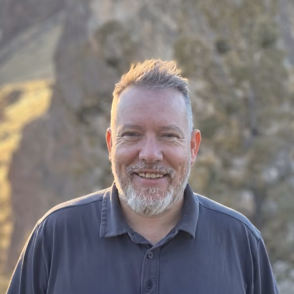 Smiling middle-aged man with short gray hair and beard wearing a dark polo shirt, standing outdoors with blurred natural rock and greenery background.