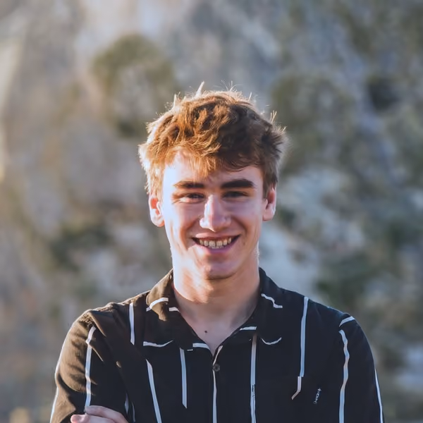Smiling young man with light brown hair wearing a dark shirt with white vertical stripes, standing outdoors with blurred nature background.