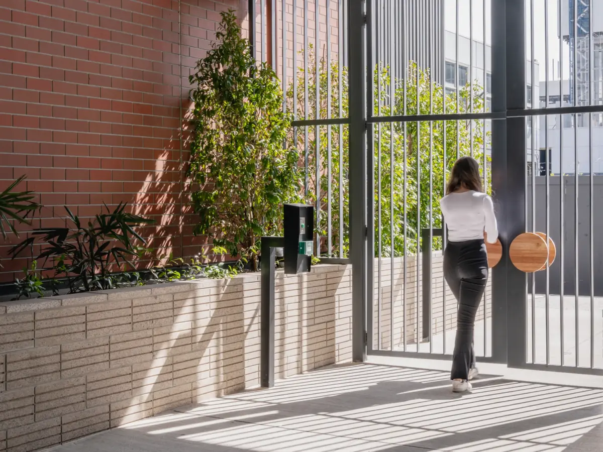 Woman in black pants and white top walking towards a metal gate with large wooden handles, next to a planter with green plants and a brick wall.