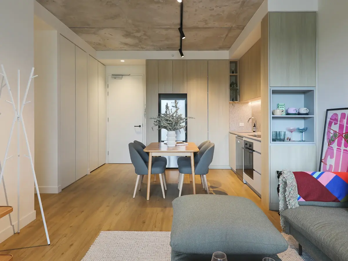 Modern apartment interior with wooden flooring, dining table with gray chairs, kitchen area with light wood cabinets, gray sofa with colorful blanket, and a white coat rack.